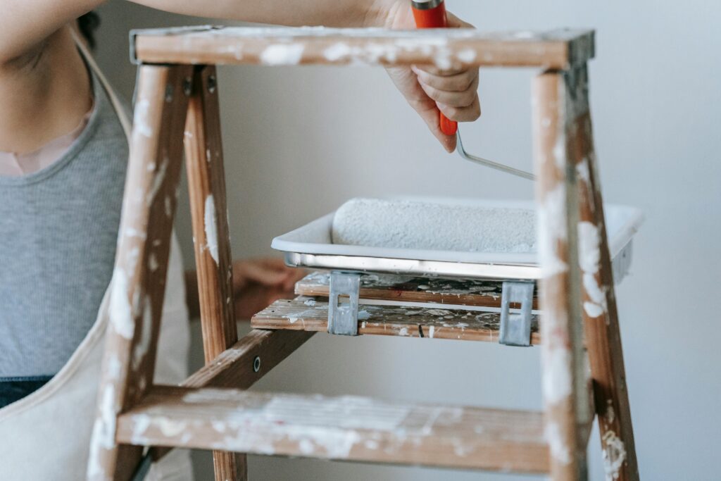 Adult woman using a roller and ladder to paint a room wall indoors.